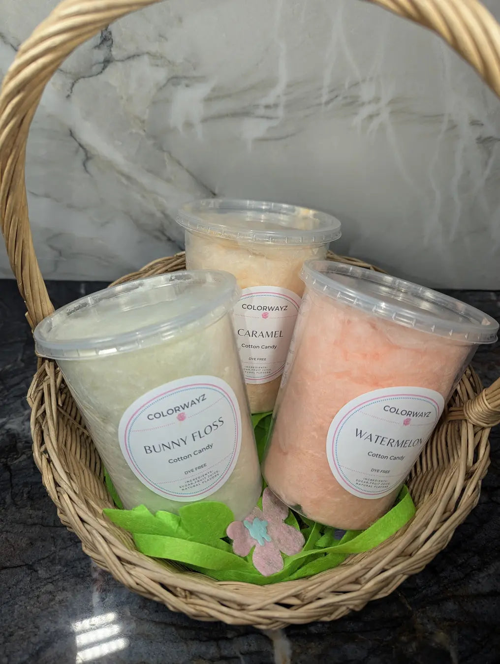 A wicker Easter basket filled with three tubs of Colorwayz cotton candy in Bunny Floss, Caramel, and Watermelon flavors, set against a marble background.