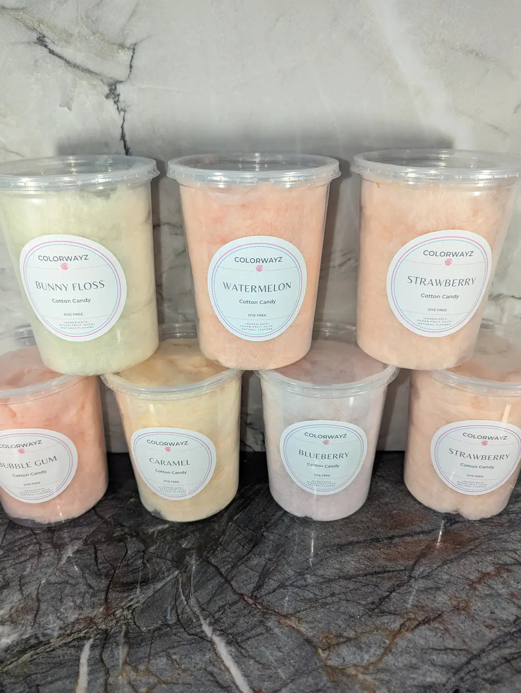 A variety of naturally colored Colorwayz cotton candy tubs in flavors like Bunny Floss, Watermelon, Strawberry, Bubble Gum, Caramel, and Blueberry, neatly displayed against a marble background.